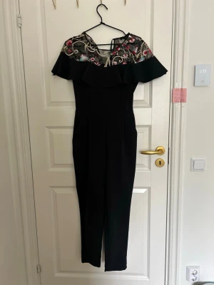 Svart jumpsuit med broderad mesh - Snygg svart jumpsuit med långa ben och off shoulder-krage. Överdelen har transparent mesh med broderade blommor i rosa, blått och grönt samt volangdetalj. Perfekt för dig som vill sticka ut med en unik och trendig look.