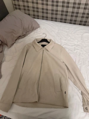 Beige manchesterjacka med dragkedja - Snygg beige jacka i manchester med dragkedja framtill och två fickor. Jackan har klassisk krage och långärmad design, perfekt för dig som gillar stilrena och enkla plagg.
