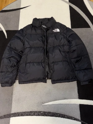 Svart dunjacka från The North Face - Säljer en svart dunjacka från The North Face med klassisk boxig passform och hög krage. Jackan har två stora fickor framtill, dragkedja och diskret logga på bröstet. Perfekt för kalla vinterdagar och riktigt skön att bära.