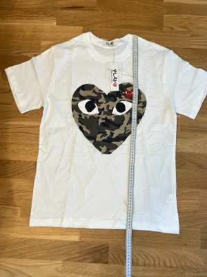 Vit t-shirt med camo-hjärta från Comme des Garçons - Säljer en vit t-shirt från Comme des Garçons Play med ett stort hjärta i camouflage-mönster och ögon på bröstet. T-shirten har korta ärmar och är gjord i mjuk bomull. Perfekt för dig som vill sticka ut med en ikonisk design.