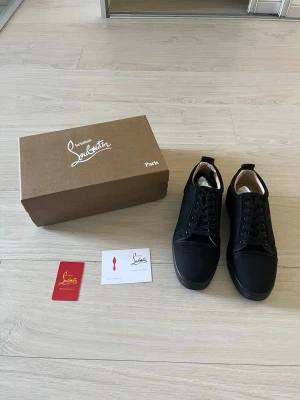 Svarta sneakers från Christian Louboutin - Säljer ett par svarta sneakers från Christian Louboutin med klassisk röd sula. Skorna har en stilren design med svarta snören och diskret logga på sidan. Materialet ser ut att vara en mix av mocka och läder, vilket ger en lyxig känsla.