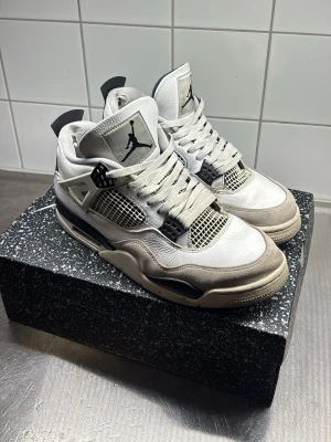  Air Jordan 4 Retro Military Black - Säljer ett par klassiska Air Jordan 4 sneakers i vitt och svart med meshdetaljer och ikonisk Jumpman-logga på plösen. Skorna har högt skaft, robust sula och coola plastdetaljer vid snörningen. Perfekta för dig som gillar streetwear och vill sticka ut.