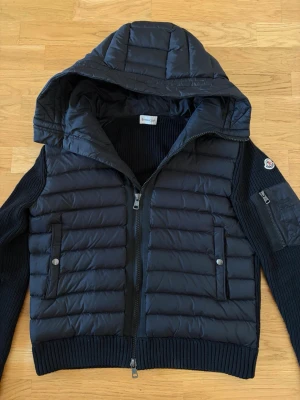 Svart pufferjacka från Moncler - Snygg svart pufferjacka från Moncler med stickade ärmar och huva. Jackan har två framfickor med tryckknappar, en ärmficka med dragkedja och ribbade muddar. Perfekt för kalla vinterdagar och har en modern, sportig look.