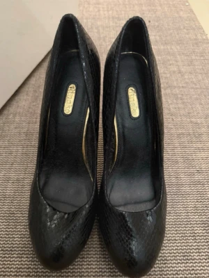 Svarta pumps med ormmönster -  svarta pumps med hög klack och ormmönstrad yta. Skorna har rundad tå och insida i svart med gulddetaljer. Perfekta för dig som vill ha en edgy och trendig look till din outfit.