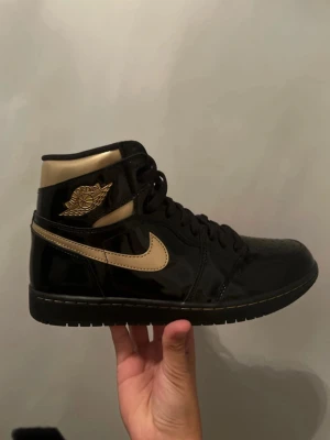 Svarta och guldiga Air Jordan 1 High - Snygga Air Jordan 1 High sneakers i svart lack med guldiga detaljer och swoosh. Skon har klassisk hög siluett, svart snörning och perforerad tåbox. Materialet är glansigt och ger en riktigt clean look. Perfekt för dig som vill sticka ut med stilrena färger.