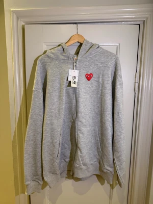 Comme des Garçons Play - Grå zip-hoodie från Comme des Garçons Play med rött hjärta-logga på bröstet. Oanvänd, storlek XL men sitter som L