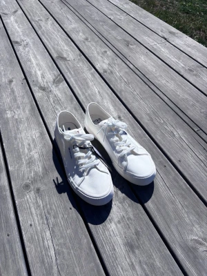 Helt Nya Tommy Hilfiger - Helt nya vita Tommy Hilfiger skor med klassisk låg profil och tygmaterial. Skorna har vita snören och en diskret logga på sidan. Perfekta för en clean och stilren look till vardagsoutfiten.