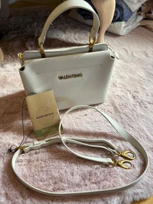 Vit handväska från Valentino - Snygg vit handväska från Valentino i strukturerat konstläder med guldfärgade metalldetaljer. Väskan har både handtag och avtagbar axelrem, samt diskret logga framtill. Perfekt för dig som vill ha en stilren och trendig accessoar. Fint använt skick. 
