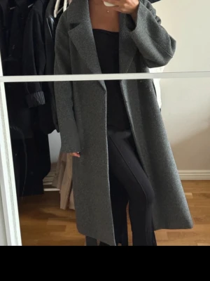 Grå lång höst kappa  - Stilren grå kappa i ullmix med rak passform och breda slag. Kappan har ett matchande knytskärp i midjan och dolda fickor i sidorna. Perfekt för kyligare dagar och ger en klassisk look till din outfit.