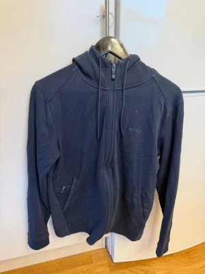 Mörkblå hoodie från Hugo Boss - Snygg mörkblå hoodie från Hugo Boss med dragkedja framtill, två fickor och justerbar huva med snören. Tillverkad i mjukt bomullsmaterial som känns skönt mot huden. Diskret Boss-logga på bröstet för en clean look. 