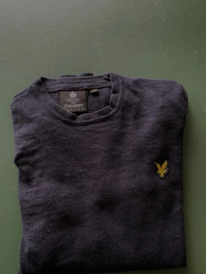 Mörkblå stickad tröja från Lyle & Scott - Mörkblå stickad tröja från Lyle o& Scott med rund halsringning och klassisk gul logga broderad på bröstet. Tröjan har ribbade muddar vid ärmslut och nederkant. Perfekt för lager-på-lager och enkeloyooo att matcha.