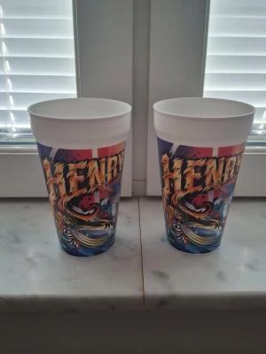 Två McDonald's plastmuggar med Henry - Säljer två vita plastmuggar från McDonald's med färgglad grafik av fotbollsspelaren Henry. Muggarna har tryck med hans namn och porträtt samt blå, röda och gula detaljer. Perfekta för samlare eller som coola dryckesglas.