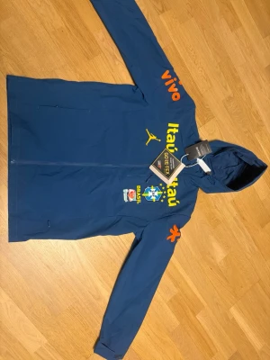 Brazil jacka jordan M - Snygg blå vindjacka med huva och dragkedja, dekorerad med Brasilien CBF-logga, gula och orangea tryck samt flera sponsorloggor. Jackan har GORE-TEX-märke och sportig look, perfekt för dig som gillar fotboll och streetwear.