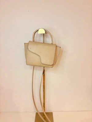 ATP Beige minihandväska med axelrem - Snygg beige minihandväska i slät läderimitation med unik asymmetrisk lockdesign. Väskan har både ett topphandtag och en lång justerbar axelrem så du kan bära den på flera sätt. Perfekt för dig som gillar stilrena och minimalistiska accessoarer. APT väska, den är använd ja, men inget slitage alls, lite fläckar på sig men som absoult går att tvätta! Säljer även här till ett bra pris, svin snygg väska ( som ni ser pga användningen 😅) men behöver bli av med den ! 