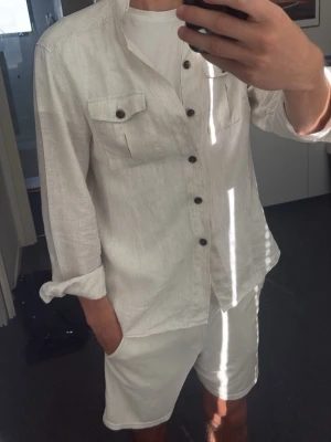 Vit overshirt John Henric - Säljer en stilren vit overshirt i linne med två bröstfickor och mörka knappar. Overshirten har lång ärm som kan vikas upp för en mer avslappnad look. Perfekt till sommaren och ger en fräsch vibe till din outfit.
