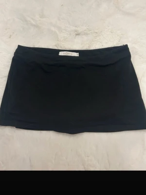 Svart skort från ALOHA - Supersnygg svart skort från ALOHA med enkel och stilren design. Skorten har en rak passform och kort längd, perfekt för en trendig och avslappnad look. Tillverkad i mjukt material som känns bekvämt hela dagen. Enda defekten är ett litet hål i sidan som inte syns pga skjortsen.