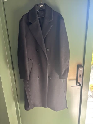Svart dubbelknäppt ullkappa Zara - Stilren svart kappa i ullmix med dubbelknäppning och bred krage. Långa ärmar och två snedställda fickor framtill. Klassisk rak passform som passar perfekt till höstens och vinterns outfits.                           Använd fåtal gånger men en defekt på ena fickan, lagad efter ett hål från fabrik