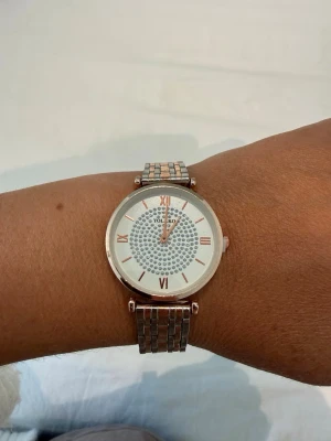 Silverfärgad klocka med stenar från YOLAKO - Stilren klocka från YOLAKO i silverfärg med glittrande stenar på urtavlan. Klockan har romerska siffror i roséguld och ett metallarmband som ger en lyxig känsla. Perfekt accessoar för att lyfta din outfit.