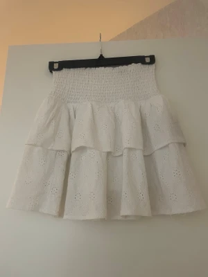 Vit volangkjol med broderade detaljer - Söt vit kortkjol med volanger och broderade blommönster. Kjolen har en smockad midja som ger en bekväm passform och ett luftigt fall. Perfekt för varma dagar och ger en romantisk vibe till din outfit.