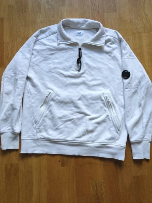 Vit half-zip hoodie från C.P. Company - Säljer en vit hoodie från C.P. Company med half-zip dragkedja och svart logodetalj på ärmen. Hoodien har två snedställda fickor med dragkedja och ribbade muddar. Tillverkad i mjukt bomullsmaterial, perfekt för en clean och stilren look. Nypris är 1900kr. Säljes för 600. Pris kan diskuteras vid snabb affär