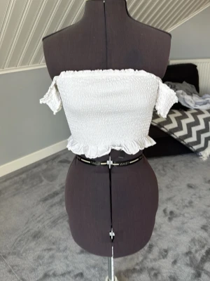 Vit offshoulder croppad topp - Säljer en söt vit offshoulder topp med smockad struktur och volangkant nedtill. Toppen är croppad och har korta ärmar som sitter nedanför axlarna. Perfekt för sommardagar och enkel att styla med jeans eller kjol.