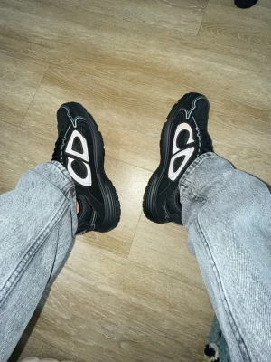 Svarta chunky sneakers med vita detaljer - Säljer ett par svarta chunky sneakers med vita och grå detaljer. Skorna har en kraftig sula och coola mönster på sidan. Perfekta för dig som gillar streetwear och vill sticka ut lite extra.