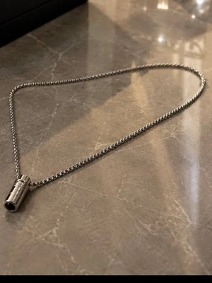 Silverfärgat halsband med cylinderhänge - Säljer ett stilrent halsband i silverfärg med en kedja i boxmodell och ett cylinderformat hänge. Halsbandet har en modern och minimalistisk design som passar till många olika stilar. Perfekt accessoar för dig som gillar enkla men snygga detaljer.