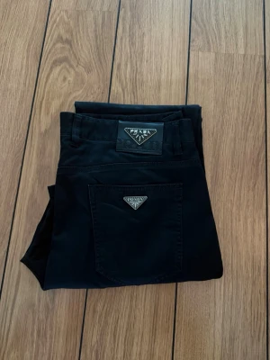 Svarta jeans från Prada - Snygga svarta jeans från Prada med klassisk femficksmodell och ikoniska metalltriangel-loggor på bakficka och framficka. Jeansen har raka ben och bälteshällor, samt silverfärgade nitar och knapp. Perfekta för en trendig och stilren look.