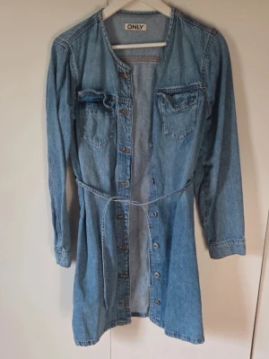 Jeansklänning med knappar från ONLY - Säljer en snygg jeansklänning från ONLY med långa ärmar, knappar framtill och två bröstfickor. Klänningen har en markerad midja med snörning och är tillverkad i klassiskt blått jeansmaterial. Perfekt för dig som gillar en avslappnad men trendig stil. 90 cm lång. Använd en gång. 