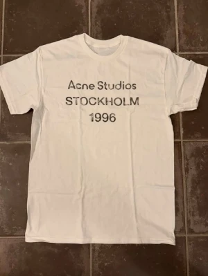 Vit Acne Studios t-shirt Stockholm - Vit t-shirt från Acne Studios med tryck 'Acne Studios STOCKHOLM 1996' på bröstet. Klassisk passform med rund hals och korta ärmar. Perfekt basic-plagg med stilren design. Är ca 182 och väger 66