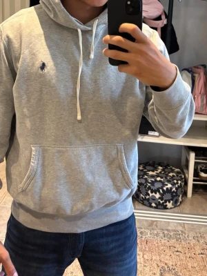 Grå hoodie från Polo Ralph Lauren - Snygg grå hoodie från Polo Ralph Lauren med klassisk huva och vita dragsnören. Har en broderad logga i mörkblått på bröstet och en stor känguruficka framtill. Tillverkad i mjukt sweatshirtmaterial som är perfekt för chill dagar.