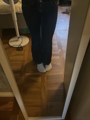 Mörkblå bootcut jeans med vita sömmar - Säljer ett par mörkblå bootcut jeans med tydliga vita kontrastsömmar och klassiska fem fickor. Jeansen har låg midja och utsvängda ben, perfekta för en trendig och avslappnad look. Materialet är denim i bomull.