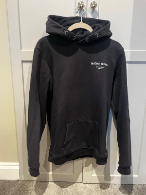 Mörk blå hoodie från Björn Borg - Snygg mörkblå hoodie från Björn Borg med vit logga och text på bröstet. Klassisk modell med huva och dragsko samt en stor magficka framtill. Tillverkad i mjukt material och har ribbade muddar vid ärmar och nederkant. Perfekt för en chill och avslappnad stil.