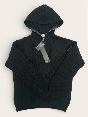 Svart Stone island hoodie med dragkedja och huva - Säljer en svart ribbstickad hoodie med dragkedja framtill och stor huva. Tröjan har en klassisk patch på ärmen och är tillverkad i ett tjockare stickat material som känns riktigt cozy. Perfekt för dig som gillar streetwear och vill ha något stilrent.