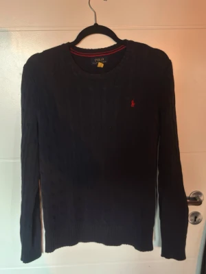 Mörkblå kabelstickad tröja Polo Ralph Lauren Strl 14-16(xs) - Mörkblå kabelstickad tröja från Polo Ralph Lauren med rund halsringning och röd broderad logga på bröstet. Långärmad modell i mjukt material, perfekt för lager på lager. Klassisk och stilren design som funkar till det mesta.