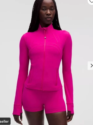 Rosa träningsset med jacka och shorts - Snyggt och figurnära träningsset från Lululemon i starkt rosa färg. Setet består av en långärmad jacka med dragkedja och höga fickor samt matchande tighta shorts. Perfekt för gymmet eller löprundan. Materialet är stretchigt och följsamt för maximal rörelsefrihet.