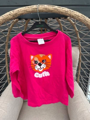 Långärmad topp strl. 130 - Rosa långärmad topp från Play Zone med ett färgglatt kattmotiv och texten 'Cutie' framtill.