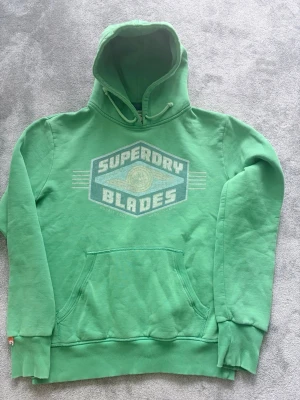 Grön Superdry Blades hoodie - Snygg grön hoodie från Superdry med stort tryck 'Superdry Blades' på bröstet. Klassisk känguruficka framtill, ribbade muddar och dragsko i huvan. Perfekt för en avslappnad och sportig stil.