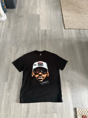 Svart t-shirt med tryck av ansikte - Cool svart t-shirt med stort tryck av ett ansikte med solglasögon och keps med texten 'USA'. T-shirten har korta ärmar och en klassisk passform. Perfekt för dig som vill sticka ut med en unik och grafisk design.