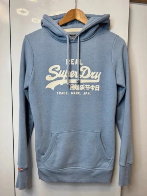 Ljusblå Superdry hoodie med tryck - Ljusblå hoodie från Superdry med vit logga och japansk text på bröstet. Klassisk känguruficka framtill, ribbade muddar och justerbar huva med dragsko. Perfekt för en avslappnad och trendig look. Mjuk bomullsblandning för extra komfort.