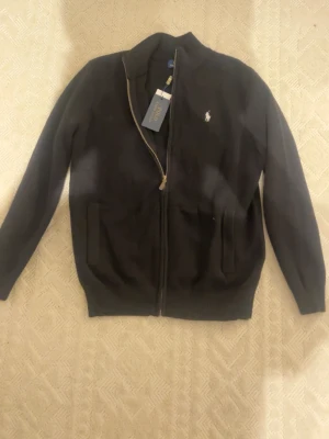 Svart ziptröja från Polo Ralph Lauren - Klassisk svart ziptröja från Polo Ralph Lauren med vit broderad logga på bröstet. Tröjan har ståkrage, två fickor framtill och ribbade muddar vid ärmslut och nederkant. Perfekt för en stilren och avslappnad look.