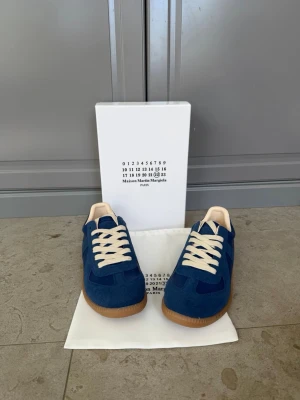 Blå Maison Margiela sneakers - Snygga blå sneakers från Maison Margiela med beige detaljer och vita skosnören. Skorna har en klassisk låg siluett och ovandel i mocka och textil, samt en brun gummisula som ger en retrovibe. Perfekta för dig som gillar stilrena och trendiga sneakers.