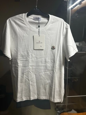 Vit t-shirt från Moncler - Säljer en klassisk vit t-shirt från Moncler med rund hals och Moncler-logga broderad på bröstet. T-shirten är enkel och stilren, perfekt till alla tillfällen. Tillverkad i mjuk bomull för skön känsla.