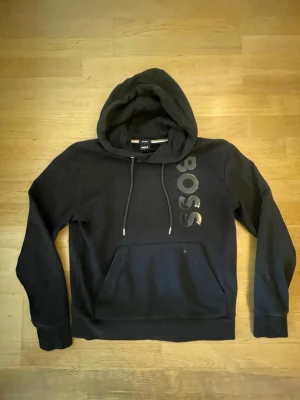 Hugo boss hoodie  - Svart hoodie från BOSS med stor logga i glänsande tryck längs bröstet. Klassisk känguruficka framtill, justerbar huva med snören och metalländar. Mjuk bomullskänsla och ribbade muddar vid ärmar och nederkant. Perfekt för en chill och stilren look.