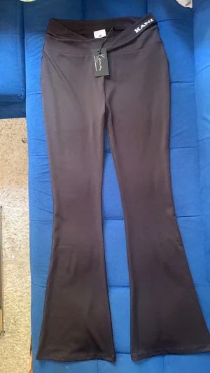 Svarta bootcut leggings från Karl Kani - Snygga svarta leggings från Karl Kani med bootcut-modell och hög midja. De har en omlott-detalj i midjan och logga på linningen. Stretchigt material som sitter skönt och formar sig efter kroppen. Perfekta för en trendig och avslappnad look.