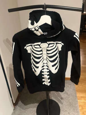 Svart skelett-hoodie från Mywear Young - Cool svart hoodie med vit skelett-tryck från Mywear Young. Perfekt för halloween eller cosplay! Tröjan har huva och ribbade muddar. 