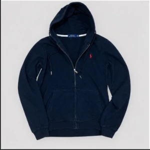Mörkblå hoodie med dragkedja - Mörkblå hoodie med dragkedja framtill och två stora fickor. Har huva med dragsko och ribbade muddar vid ärmar och nederkant. Liten röd logga broderad på bröstet. Perfekt för en avslappnad och stilren look.