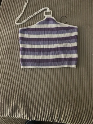 Lila och vit randig croppad topp - Säljer en ribbad, croppad topp med halterneck i lila och vitt. Toppen har breda ränder och är ärmlös, perfekt för sommaren. Sitter tight och ger en fräsch vibe till din outfit.
