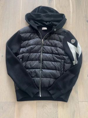 Svart Moncler Cardigan med stort M logga - Snygg svart hoodie med vadderad framsida och stickade ärmar. Dragkedja framtill, huva och två fickor. Ena ärmen har en vit blixtdetalj och ett märke på axeln. Perfekt för en avslappnad stil.