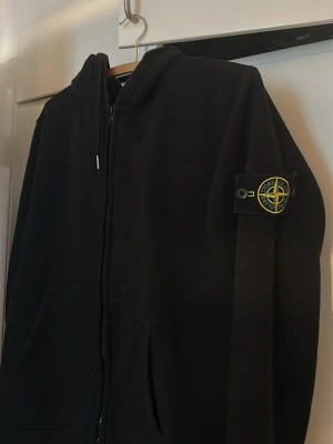 Svart hoodie från Stone Island - Säljer en svart hoodie från Stone Island med dragkedja framtill och klassisk logopatch på vänster ärm. Hoodien har huva och kängurufickor. Perfekt för dig som gillar streetwear och vill ha en stilren look. Vid snabb affär kan pris diskuteras 🌟